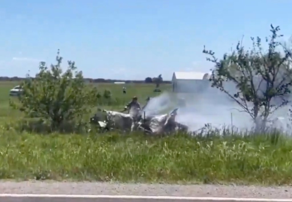 Wreckage of a plane smoking in a grassy field with emergency responders nearby.