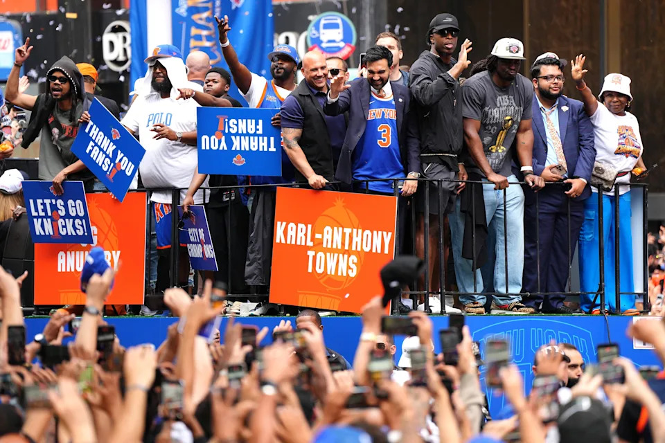 Group of people, including athletes, on a parade float holding signs supporting a sports team, surrounded by a cheering crowd taking photos