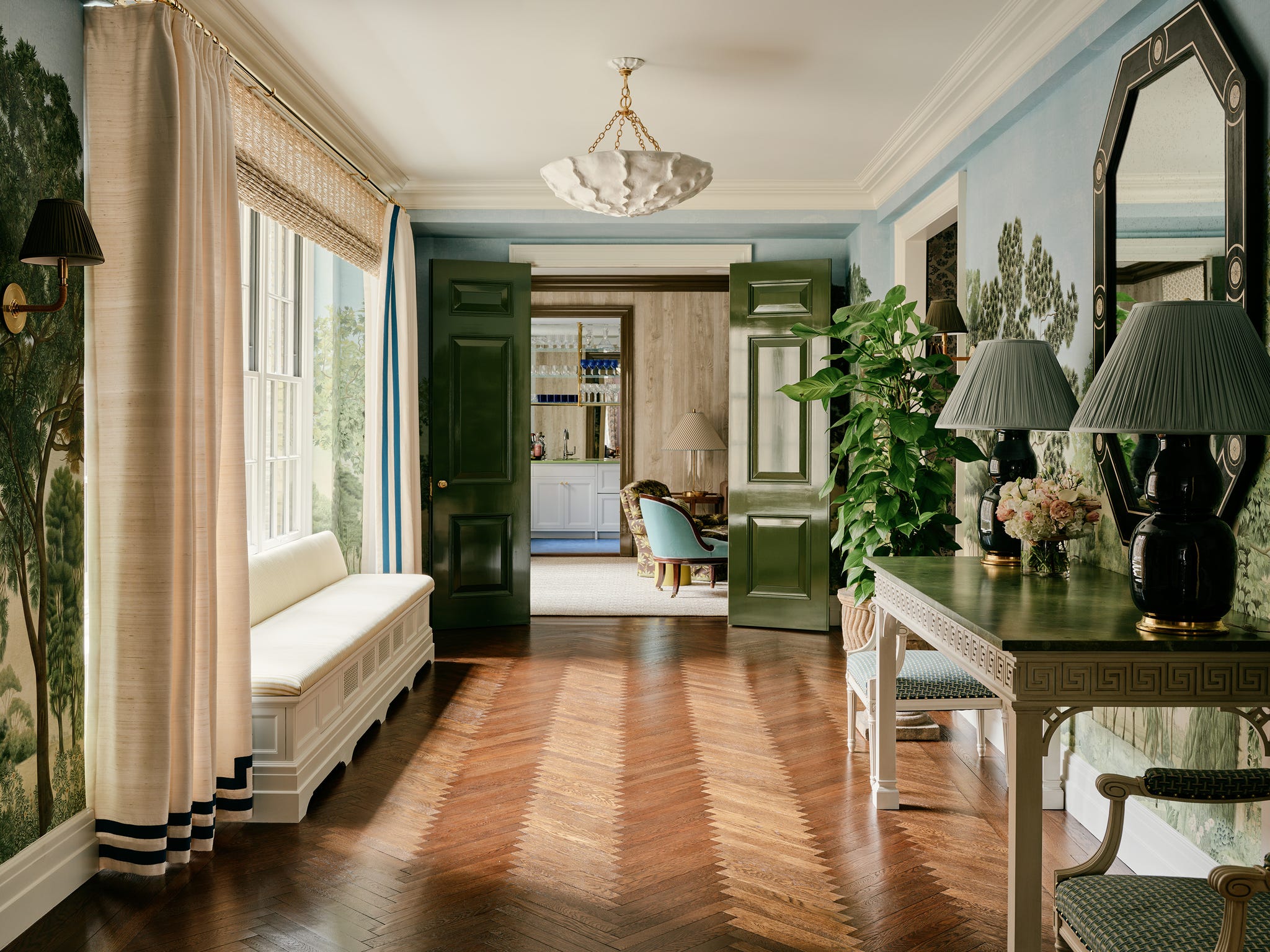 Elegant hallway with green door, decorative table, lamps, and large mirror reflecting trees.