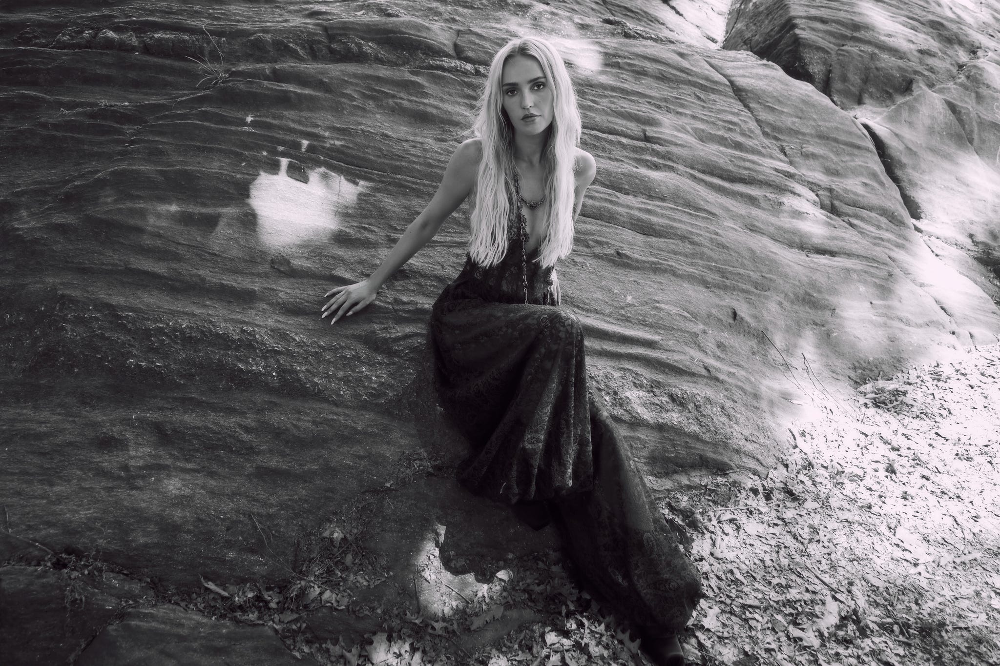 Person in a flowing dress seated on a large rock outdoors in black and white.
