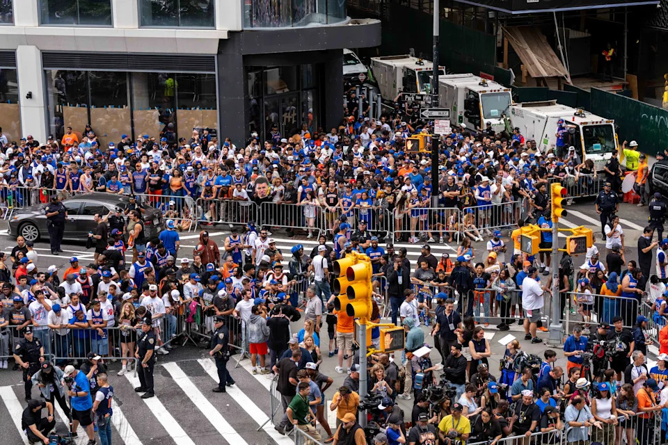 Large crowd gathered at a city intersection, many wearing sports jerseys and hats. Barriers and police are present, directing and controlling the crowd