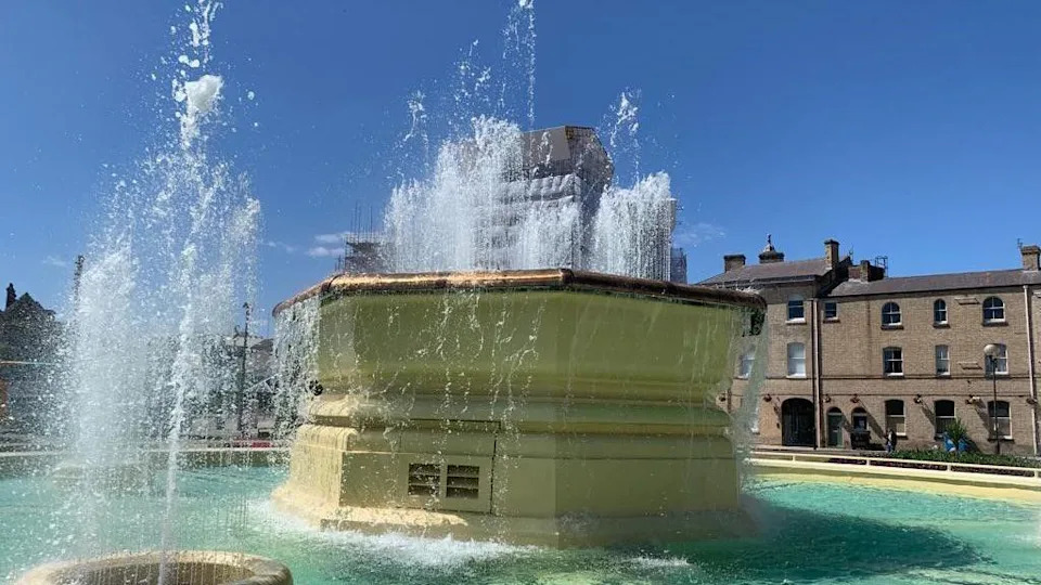 A large fountain, painted yellow, spurting out blue water under blue skies.