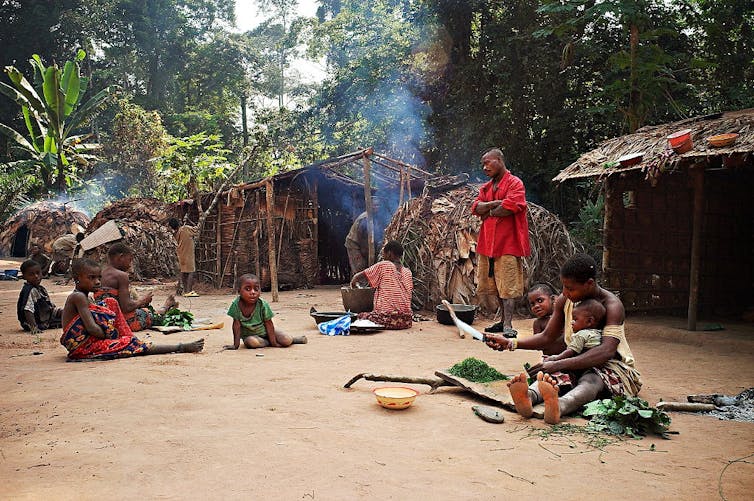 Children sit on the ground in various poses. Some cook and help with food prep. A man in a red shirt stands with his arms folded, watching.
