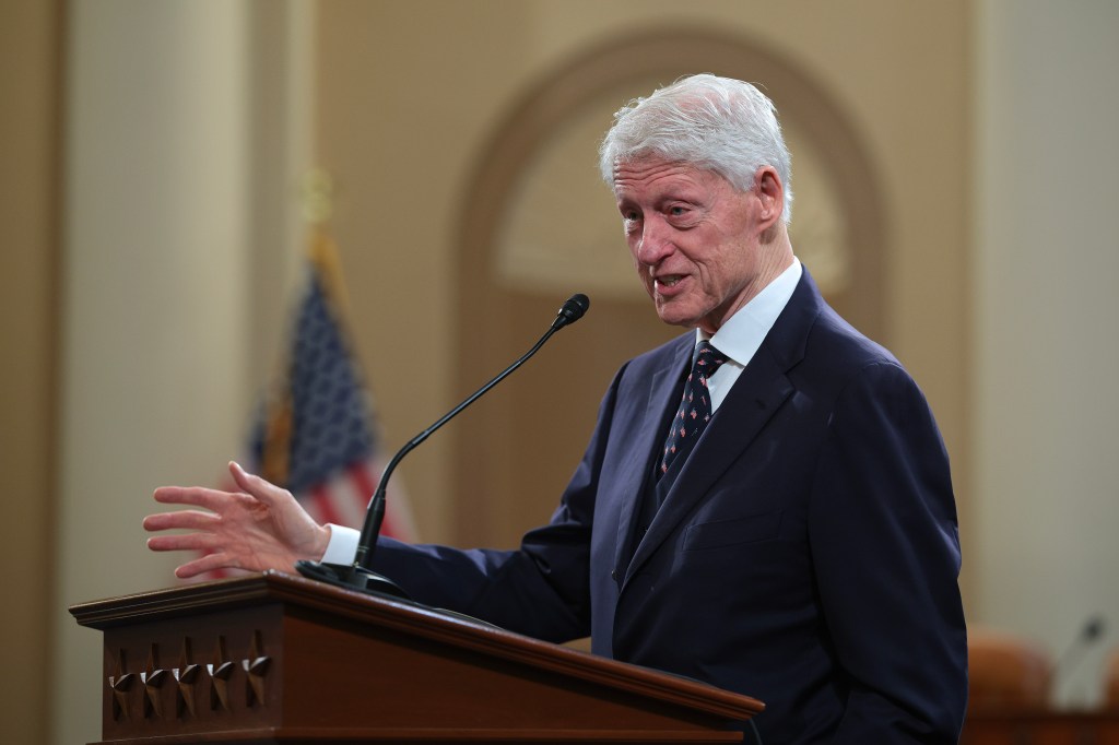 Former President Bill Clinton speaking at a podium with a microphone, gesturing with his left hand.
