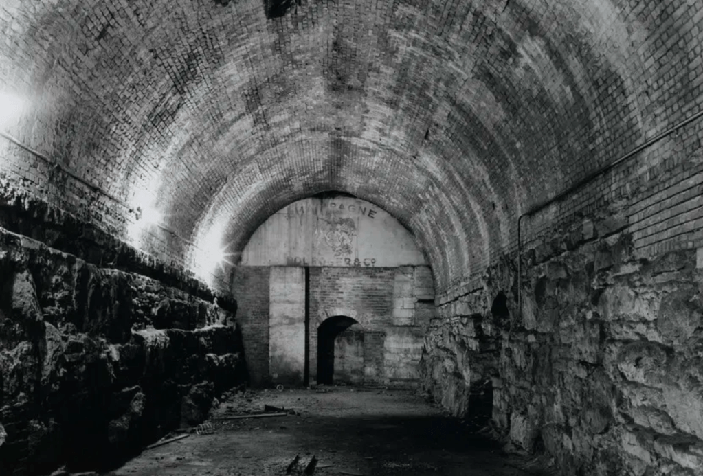 A dimly lit underground tunnel featuring arched brick walls and stone foundations.