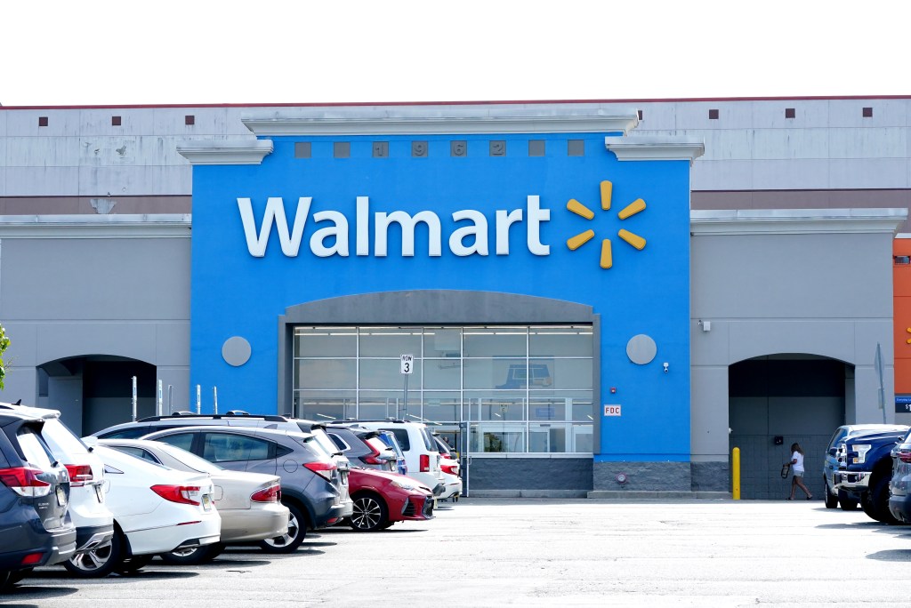 The front of a Walmart store with its blue and gray facade, a large "Walmart" sign, and cars parked in the foreground.