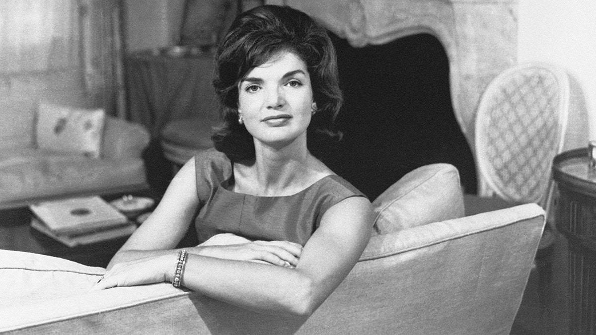 Jacqueline Kennedy seated leaning on the back of a couch in a living room