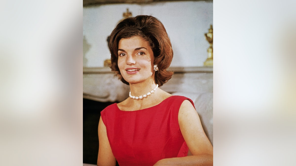 Jacqueline Kennedy wearing a sleeveless red dress with pearls standing in her Georgetown home.