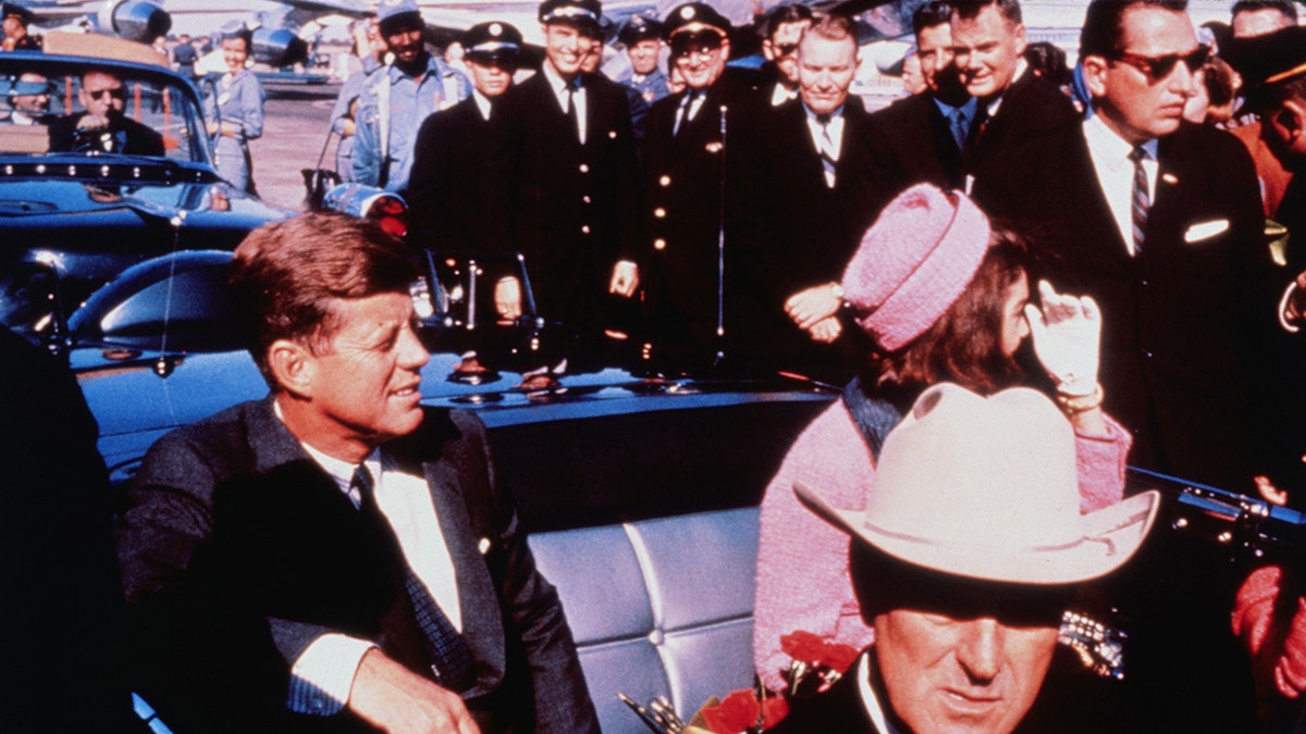 Texas Governor John Connally adjusts his tie as President John F. Kennedy and First Lady Jacqueline Kennedy sit in rear seats of automobile.