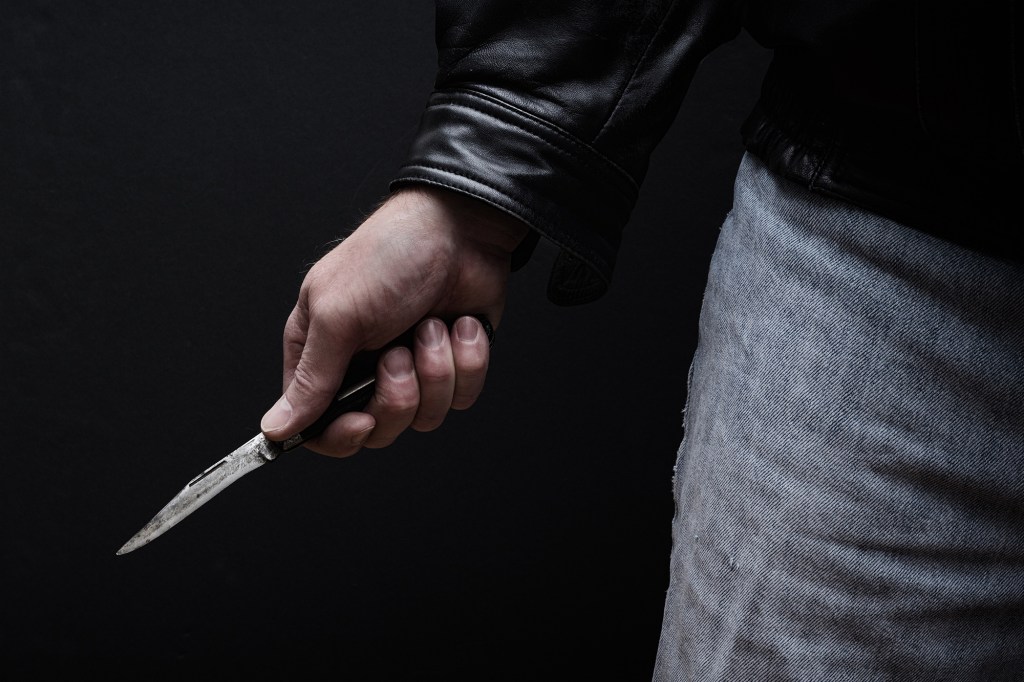 Man in a black leather jacket and jeans holding a knife with a black handle.