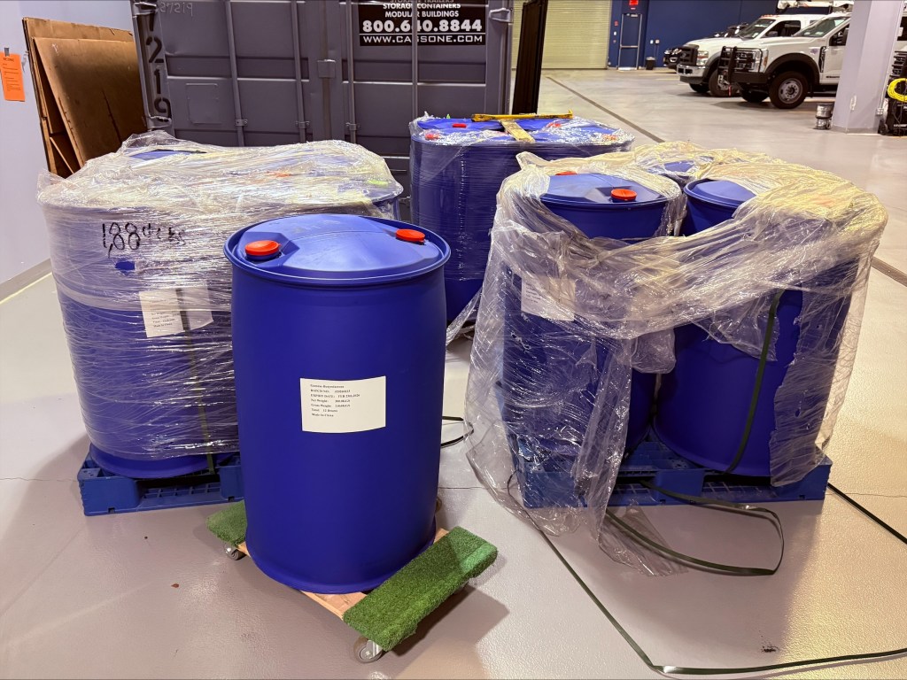 Blue drums of Gamma-Butyrolactone (GBL), wrapped in plastic, in a warehouse.