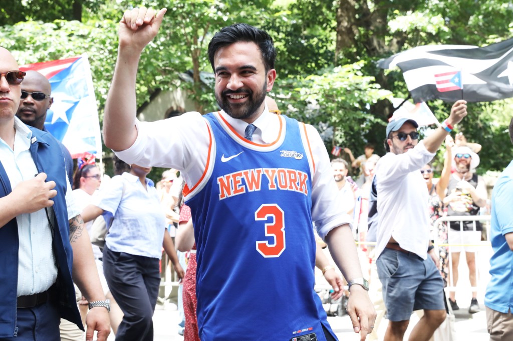 Mayor Zohran Mamdani, smiling, wearing a blue and orange "New York 3" jersey over a white shirt and tie, waves his right arm in the air during the Puerto Rican Annual parade.