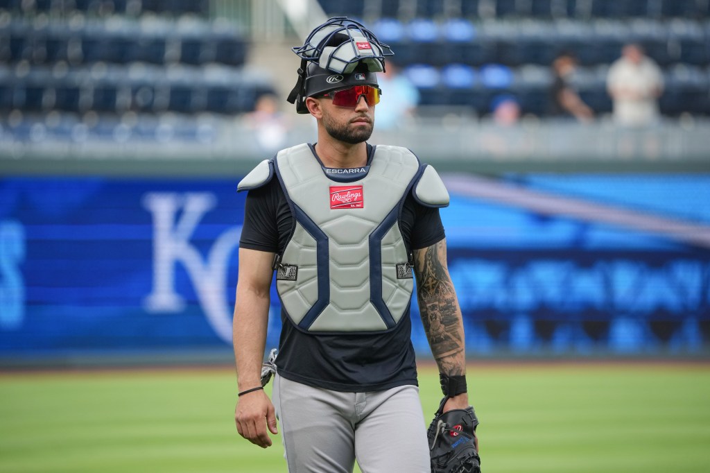 New York Yankees catcher J.C. Escarra wearing full catching gear, including a mask perched on his head, red sunglasses, a chest protector, and a glove, walking on the field.