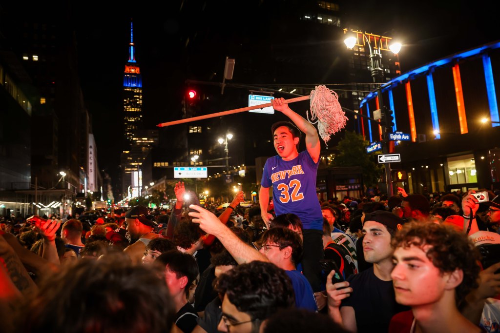A Knicks fan in a jersey holding a mop over his head in a street full of celebrating fans at night.