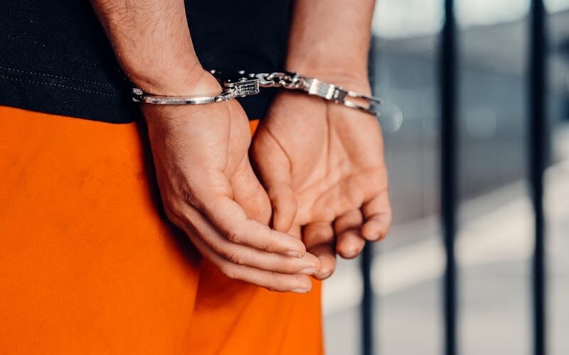 A person in orange jail attire stands with their hands cuffed behind their back.
