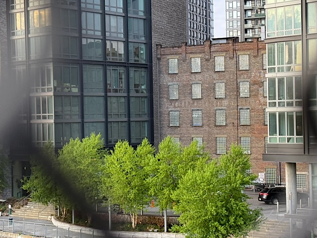 A view of urban buildings, featuring a mix of modern glass architecture and an older brick structure, surrounded by greenery near a waterfront.