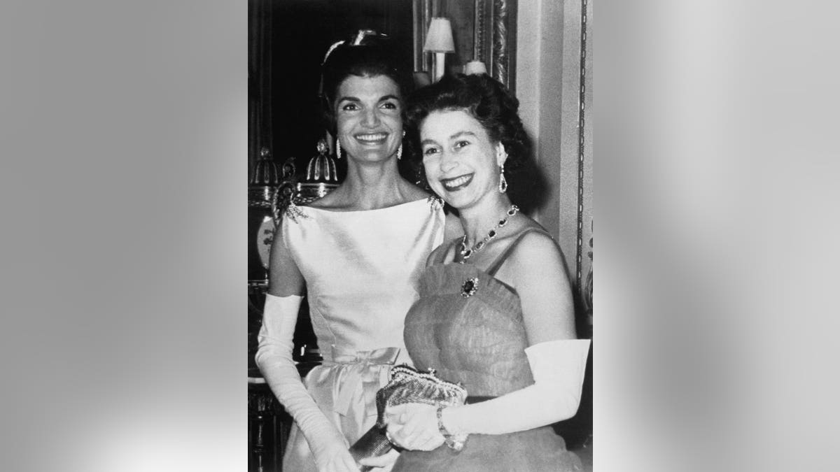 Queen Elizabeth standing next to Mrs. Jacqueline Kennedy, both smiling at Buckingham Palace.