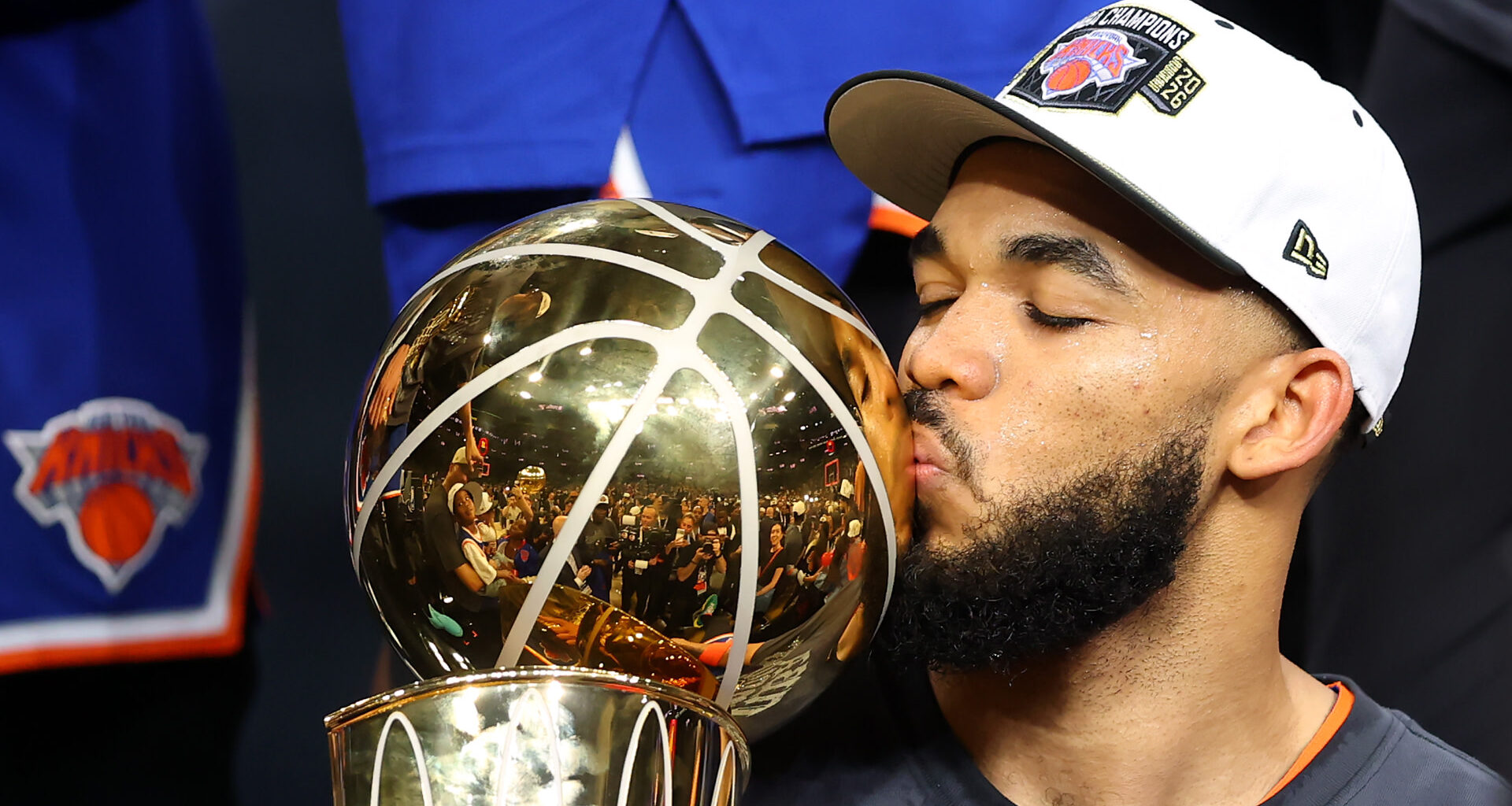 Karl-Anthony Towns kisses a trophy, wearing a cap with a New York Knicks logo. The trophy is gold and shiny, reflecting the crowd.