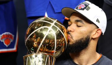 Karl-Anthony Towns kisses a trophy, wearing a cap with a New York Knicks logo. The trophy is gold and shiny, reflecting the crowd.
