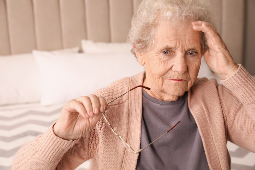 Senior woman in her bedroom, holding her glasses with one hand and her head with the other