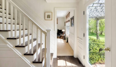 Bright entryway with white walls, wooden stairs, and a glass front door letting in sunlight. A room with beige carpet and a window is visible to the left, and greenery can be seen outside through the open door.
