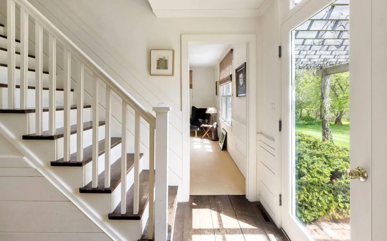 Bright entryway with white walls, wooden stairs, and a glass front door letting in sunlight. A room with beige carpet and a window is visible to the left, and greenery can be seen outside through the open door.