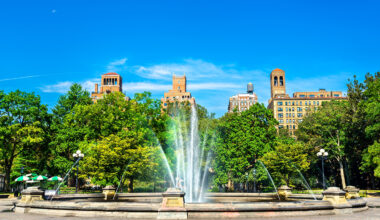 A large fountain sprays water upwards in the center of a park, surrounded by green trees. Behind the trees, historic buildings with red rooftops evoke classic NYC residential rental charm against a clear blue sky.