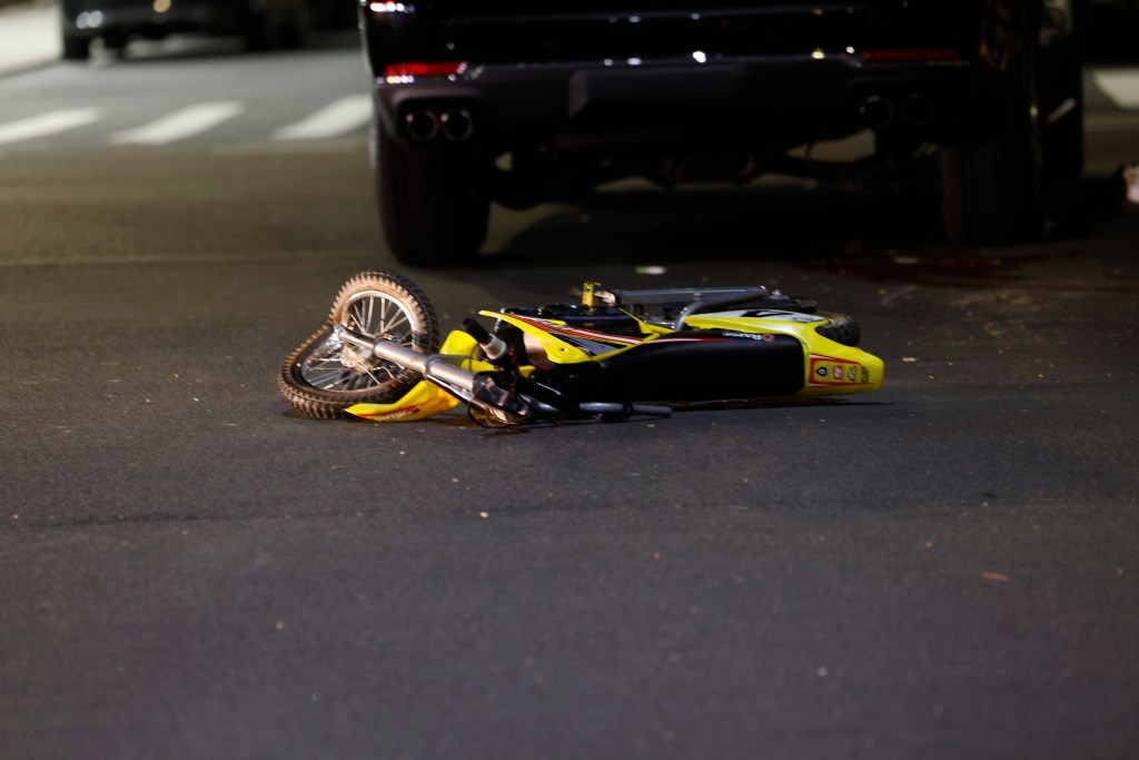 A yellow child's bike lies on its side on a dark asphalt road in front of the back of a dark car.