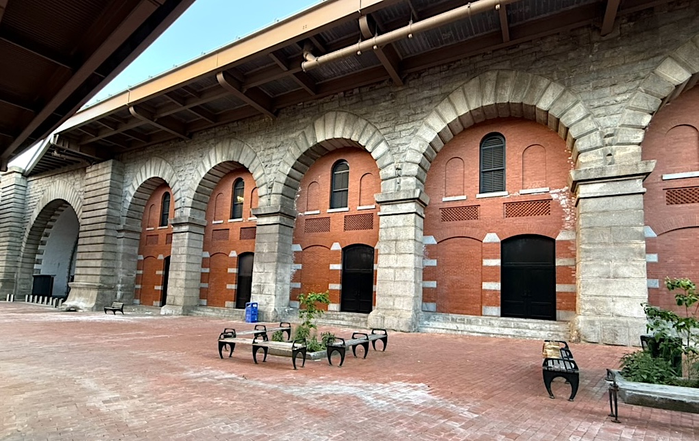 Exterior of an old brick building featuring arched windows and doors with a stone facade, set in a courtyard with benches and greenery.