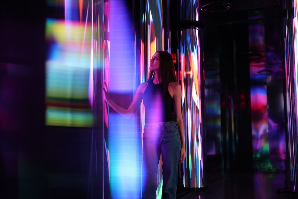 Woman standing amidst vibrant, colorful lights reflecting on glass walls - Edge NYC's Prism