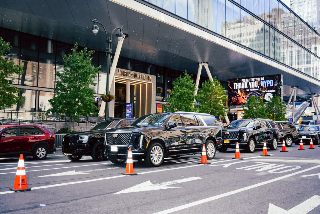 Black SUVs are double-parked outside Madison Square Garden next to a line of orange plastic cones. A digital screen, background, says “For All That You Do Thank You, NYPD.” 