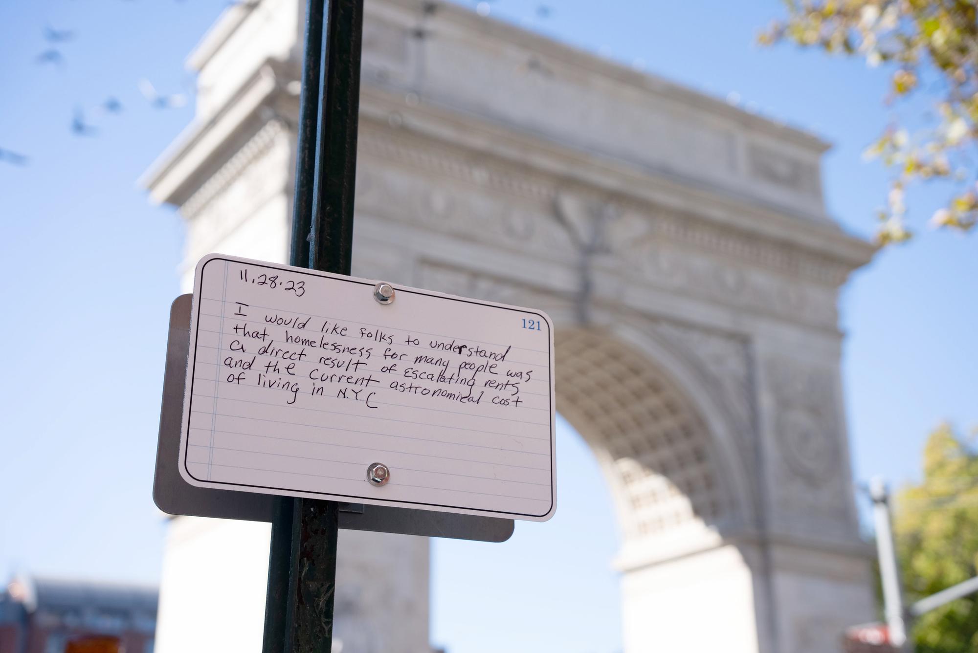 A handwritten note affixed to a pole reads: "11.28.23 I would like folks to understand that homelessness for many people was a direct result of escalating rents and the current astronomical cost of living in NYC." In the background, an archway structure is visible against a blue sky, with several birds flying above.