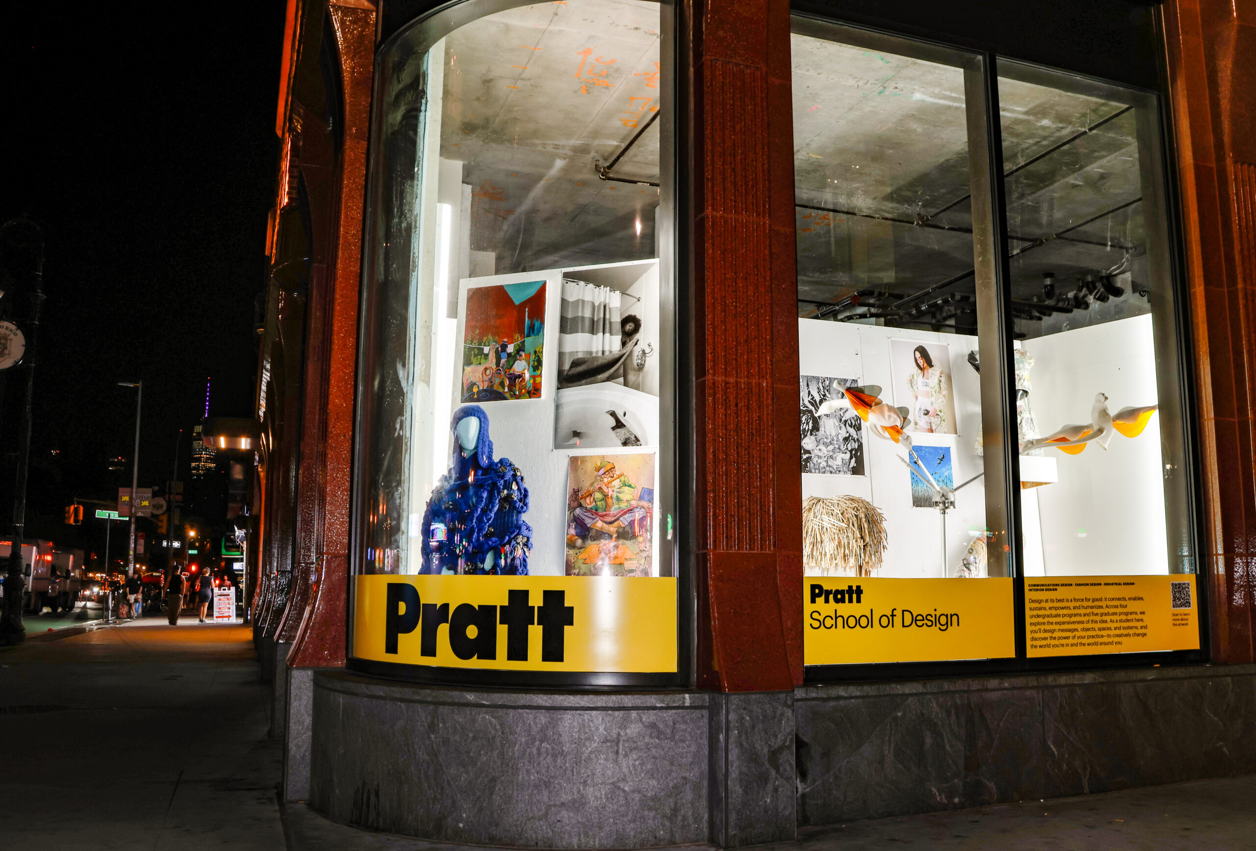 A brightly lit storefront displays artwork and installations at night. The window features a blue artistic figure, various framed images, and colorful decorations. Prominently displayed is the name 