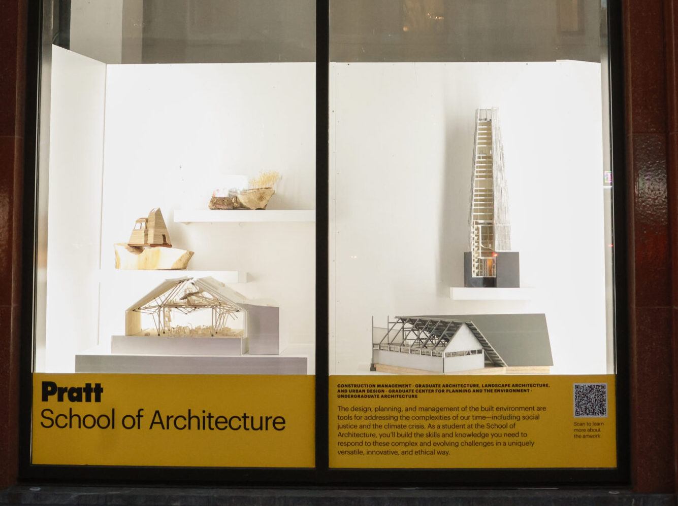 A window display features architectural models showcasing various designs. On the left, a small wooden structure is visible, while adjacent are models that include a larger, angular building and a house-like structure with a framed roof. The bottom of the display contains text about the Pratt School of Architecture, emphasizing its focus on design, planning, and management of built environments, along with a QR code for more information.