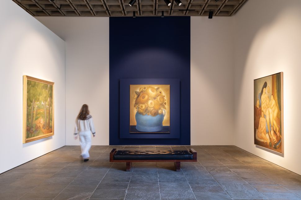A pair of art handlers installs a wide, dark-toned painting of overlapping figures and vivid geometric shapes on a freestanding museum wall.