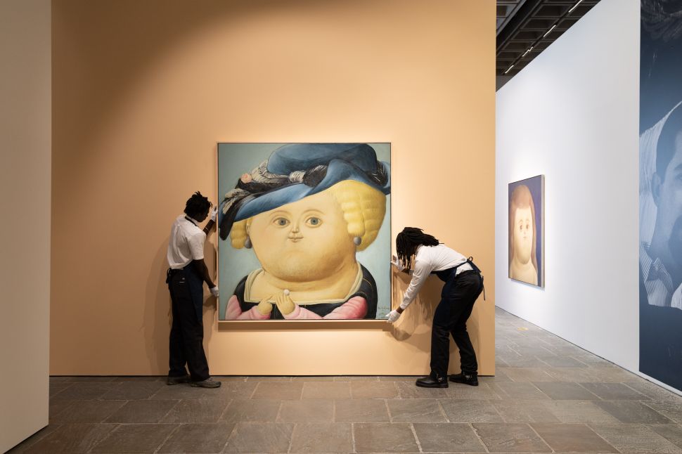 A pair of art handlers installs a large square portrait of a round-faced woman wearing a dark blue hat against a pale peach wall.