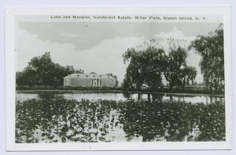 Black and white photo of a serene lake with a mansion in the background.