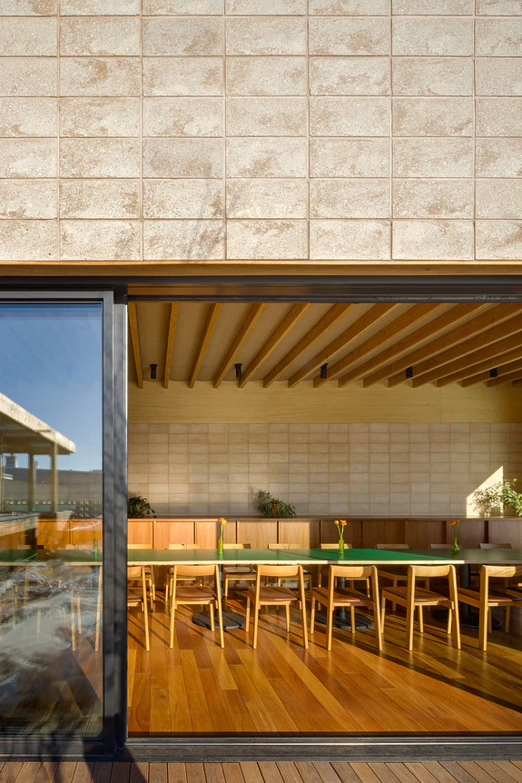 Sunlit modern restaurant interior with wooden floors, tables, and chairs, viewed through large glass doors and windows.