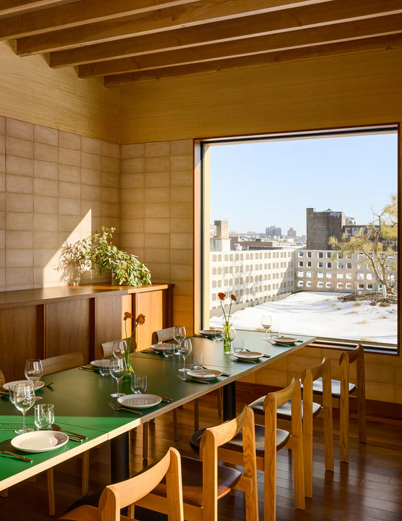 Modern dining room with a green table, wooden chairs, and a large sunlit window overlooking a snowy urban landscape.