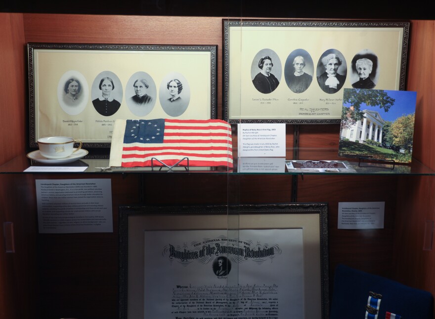 Photos of the original members of the Rochester-area Daughters of the American Revolution are displayed in a case as part of an exhibit exploring Rochester's connections to the American Revolution and its legacy. The exhibit features original artifacts from the New York State Museum and local collections and is presented by the Office of Rochester & Monroe County History, the Irondequoit Chapter of the Daughters of the American Revolution, and the Sons of the American Revolution.