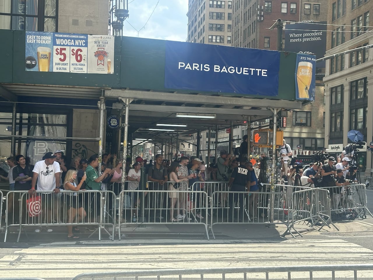 People are seen gathering behind metal barricades on a city street corner on a sunny day.