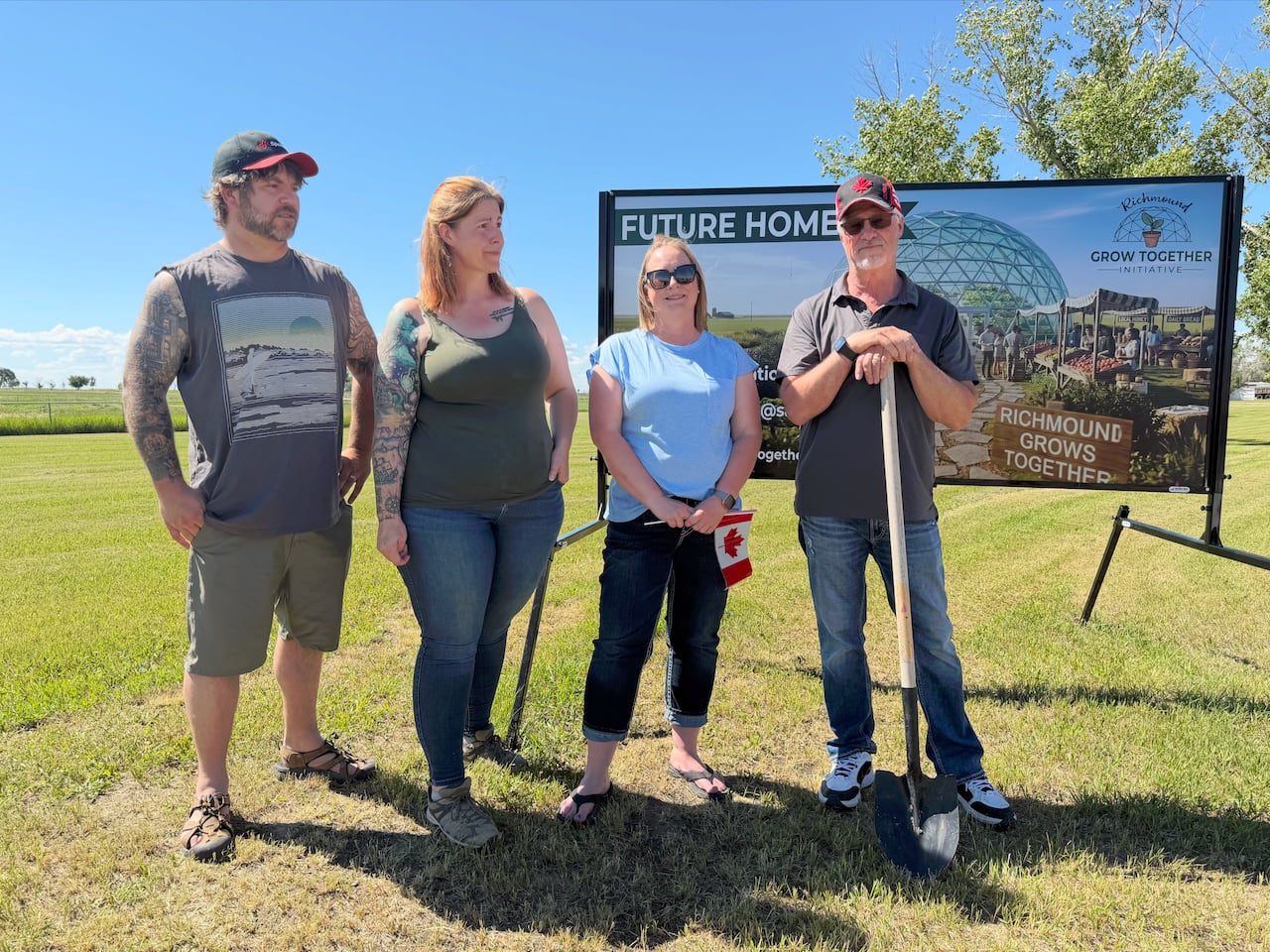 Four adults stand in a field in front of a sign for a proposed greenhouse. One of them has a shovel.
