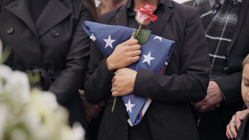 Funeral, graveyard and woman with American flag for veteran for respect, ceremony and memorial service. Family, depression and sad people with flower in cemetery for military, army and soldier hero