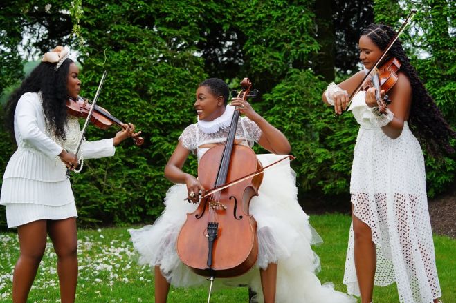 Three women in white dresses playing string instruments in a lush, green outdoor setting.