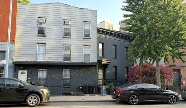 A street view showing two buildings: a gray multi-story building with multiple windows and an adjacent black building with distinct architectural features, surrounded by parked cars and greenery.