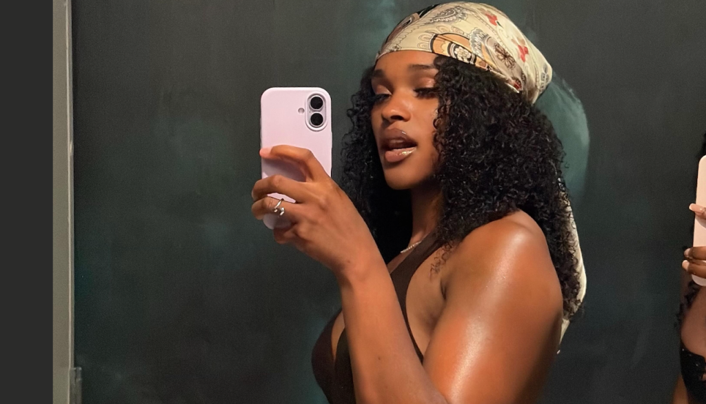 A young woman with curly hair taking a selfie on a smartphone against a teal background.