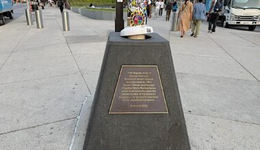 A historical plaque marking the location of the former Ebbets Field in Brooklyn, featuring a flagpole and colorful artwork on top, surrounded by pedestrians and city scenery.