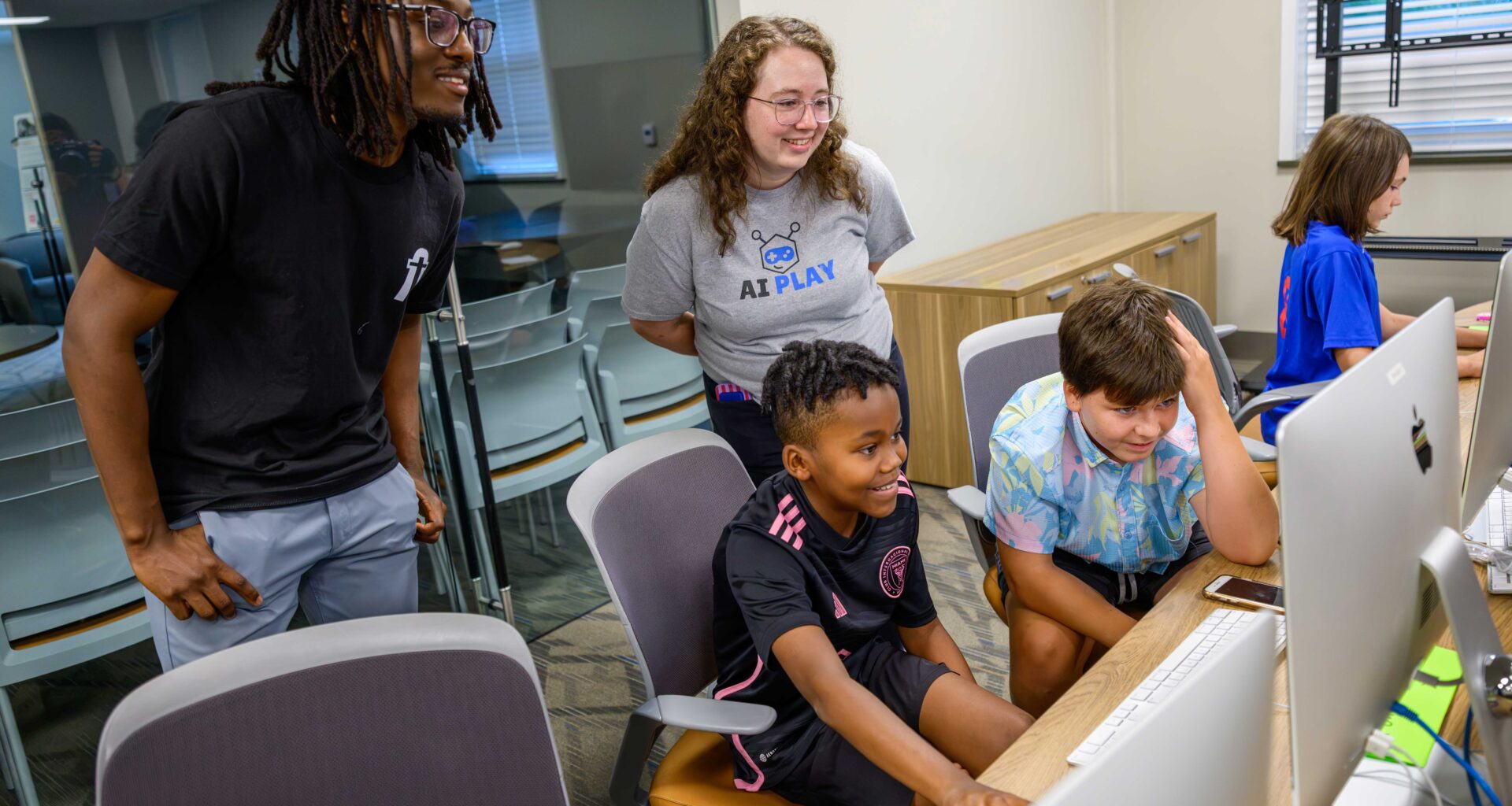 Man with black shirt and glasses and woman with glass and a tee shirt with the words AI Play peer over two boys sitting in front of computers looking at their screens.