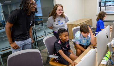 Man with black shirt and glasses and woman with glass and a tee shirt with the words AI Play peer over two boys sitting in front of computers looking at their screens.