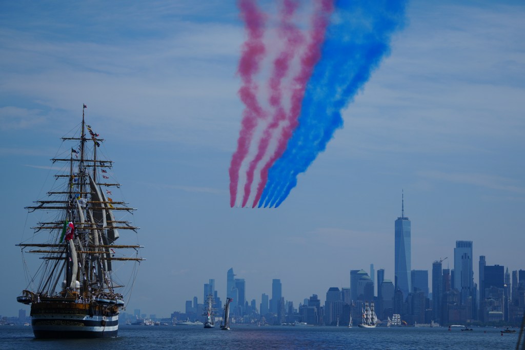Tall ships and the New York City skyline as jets fly overhead leaving red and blue smoke trails.